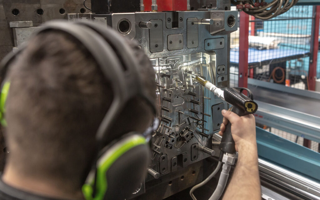 Engineer doing maintenance on a injection mold for plastic components, industrial and automotive concept En person som bär hörlurar arbetar på en metallisk maskindel med hjälp av ett handhållet verktyg i en industriell miljö.